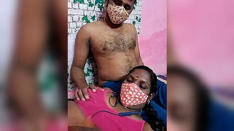 Media: Video of a shirtless, bearded Indian man wearing a face mask, sitting on a bed with a woman in a pink top and face mask, in a room with a green and white wall.