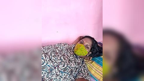 Media: Video of a young girl lying on a patterned bedspread, wearing a colorful mask, in a pink-walled room. Her dark hair is tousled, and she appears relaxed.