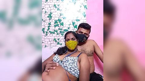 Media: Video of a pregnant woman with dark skin, wearing a yellow mask and floral bra, sitting next to a shirtless man with dark skin and a black mask, against a pink and white wall.