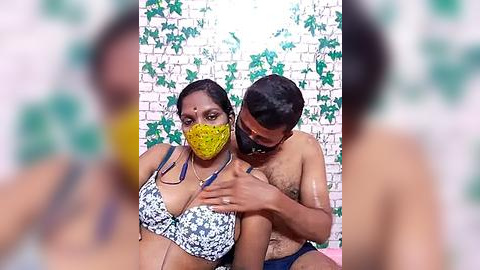 Media: Video of a topless Black man with a beard and a Black woman wearing a yellow face mask, sitting against a white brick wall with green ivy.