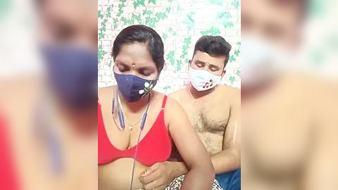 Media: A video of two people, a woman in a red bra and blue mask, and a man shirtless with a white mask, sitting on a bed with green and white patterned sheets.