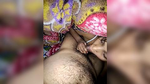 Media: Video of a close-up intimate scene: a woman with medium brown skin and a colorful patterned cloth covering her head and shoulders, performing oral sex on a man with a hairy chest.