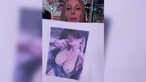 Media: Video of a blonde woman in a revealing black lace bra, holding a photo of herself in a similar outfit, with a colorful tattooed face in the background.