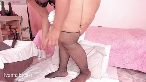 Media: Video of a light-skinned woman with large breasts, wearing sheer black stockings, adjusting her garter belt in a pink-tinted bedroom with pastel decor.