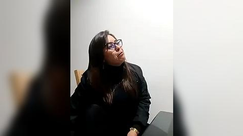 Media: Video of a woman with long dark hair and glasses, wearing a black jacket, seated in a minimalist room with white walls, partially obscured by a person's arm in the foreground.