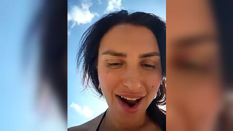 Media: Video of a woman with dark hair, light skin, and a slightly open mouth, captured outdoors with a blue sky and fluffy clouds in the background.