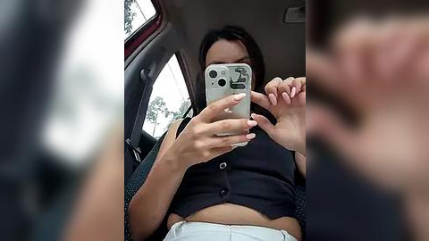 Media: A video captures a young woman with light brown skin and dark hair, wearing a black crop top and high-waisted white shorts, taking a selfie in a car.