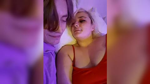 Media: Video of a blonde woman in a red tank top, lying on a bed with a man leaning over her, smiling. Background is blurred, bathed in soft, purple lighting.