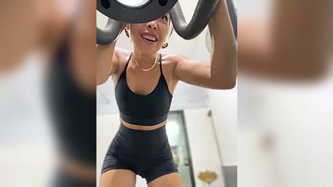 Media: Video of a fit woman with medium skin tone, wearing a black sports bra and shorts, doing a squat with a dumbbell in a gym.