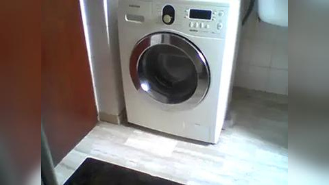 Media: Video of a modern, white front-loading washing machine with a digital control panel and glass door, positioned against a wall with white tiled floor and a partially visible toilet in the background.
