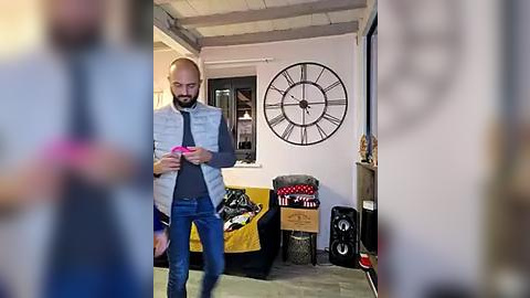 Media: Video of a bearded, bald, middle-aged man in a grey vest and blue jeans, holding a pink smartphone, in a cozy, rustic living room with white walls, a large clock, and a colorful, patterned chair.