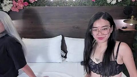 Media: Video of an Asian woman with long black hair, wearing glasses and a black lace bra, smiling in a hotel room with white pillows and a wooden headboard adorned with floral decorations.