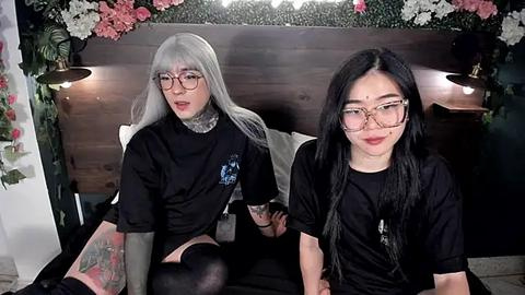 Media: Video of two women with long hair, one in a black turtleneck, the other in a black tee, sitting on a bed with floral decorations, wearing glasses.