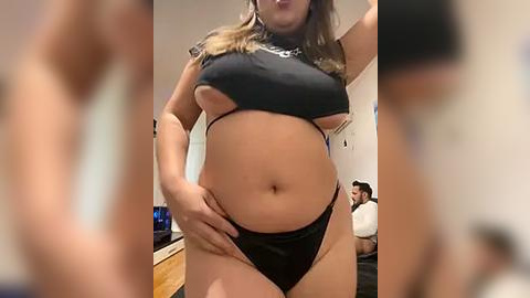 Media: Video of a plus-sized woman with light skin, wearing a black crop top and black thong, standing indoors.