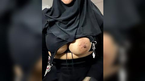 Media: Video of a woman in a black burqa, revealing her large, bruised breasts and nipples through the fabric. She wears black leggings. Background is blurred, focusing on the woman's torso.