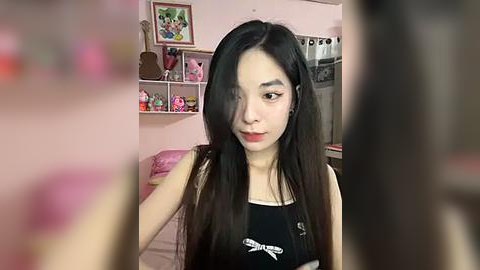 Media: Video of an Asian woman with long black hair, wearing a black tank top, sitting in a pink room with shelves filled with toys and framed pictures.