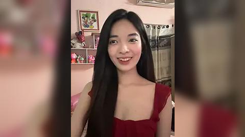 Media: A video of a young Asian woman with long black hair, smiling, wearing a red sleeveless top, standing in a cozy, pink-painted room with framed artwork and shelves filled with decorative items.