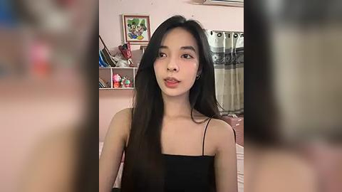 Media: Video of a young Asian woman with long black hair, wearing a black spaghetti-strap top, in a bedroom with pink walls, a framed photo, shelves, and curtains.