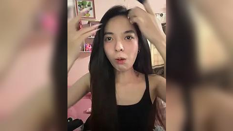 Media: Video of an Asian woman with long black hair, wearing a black tank top, adjusting her hair in a pink room with framed artwork and toys.