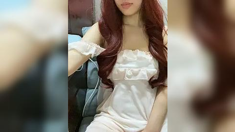 Media: Video of a young Asian woman with long, wavy, auburn hair, wearing a white, off-shoulder nightgown, seated in a dimly-lit room. Background is blurred, focusing on her face and upper body.