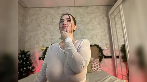 Media: A video of a fair-skinned woman with blonde hair, wearing a tight white top, standing in a cozy, dimly lit bedroom with Christmas decorations, a tree, and a bed.