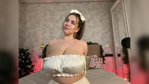 Media: Video of a curvy, fair-skinned woman with large breasts in a white ruffled crop top and high-waisted skirt, standing in a cozy, softly lit bedroom with floral wallpaper, a bed, and Christmas decorations.