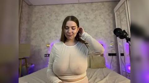 Media: Video of a young Caucasian woman with long brown hair, wearing a tight white crop top, sitting on a bed in a dimly lit room with floral wallpaper.