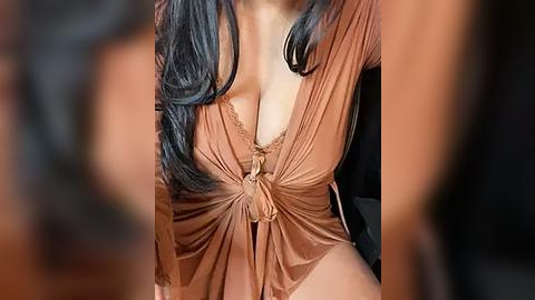 Media: Video of a woman with long, dark hair, wearing a low-cut, brown satin robe, revealing ample cleavage, and sitting in a dimly lit room.