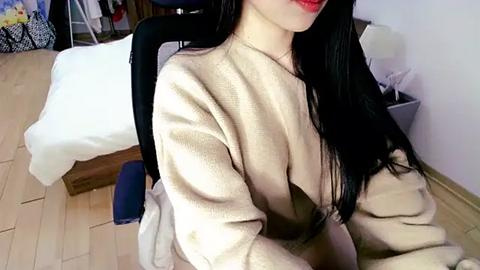 Media: A video of an Asian woman with long black hair, wearing a beige cardigan, sitting on a blue gaming chair in a bedroom with a white bed, wooden floor, and a lamp.