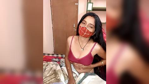 Media: Video of a young woman with medium skin tone, long black hair, wearing a red mask, pink bra, and white pants, seated on a bed in a modest room with wooden furniture and a cluttered background.