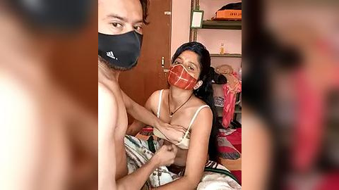 Media: Video of a topless man with a mask and a woman in a plaid face mask, bra exposed, in a messy room.