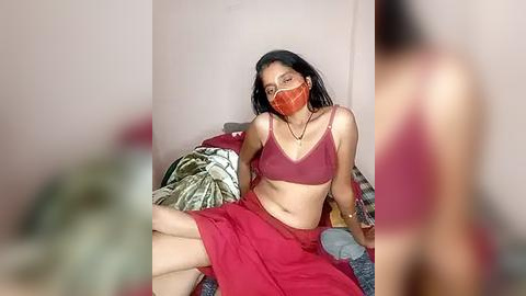 Media: Video of a South Asian woman with medium brown skin, wearing a red bralette, matching skirt, and orange mask, seated on a bed with patterned pillows in a dimly lit room.