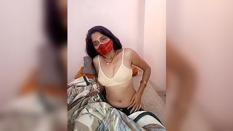 Media: Video of a woman with long black hair, wearing a red mask and beige bra, sitting on a bed with messy sheets, in a dimly lit room with white walls.