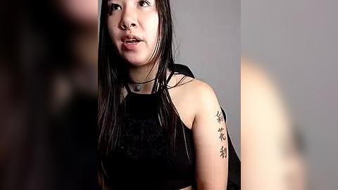 Media: Video of an Asian woman with long black hair, wearing a black sleeveless top, and a choker necklace, speaking with a man blurred in the background.