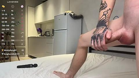 Media: A video shows a nude man with tattoos, holding his erect penis, in a modern kitchen with white cabinets, a black and white tiled floor, and a gray fridge.