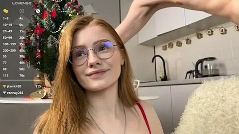 Media: A young Caucasian woman with long red hair, wearing glasses and a red top, smiles in a modern kitchen with a Christmas tree and kitchen utensils.