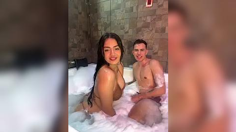 Media: Video of a young woman with long black hair, wet from a bath, smiling, and a young man in the background, both in a beige tiled bathroom.