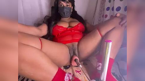 Media: Video of a curvy, dark-skinned woman with long black hair, wearing a red lace bra and mask, lying on a bed with legs spread, surrounded by vibrators, in a dimly lit room.