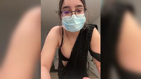 Media: Video of a young Asian woman with glasses, wearing a blue surgical mask and a black sleeveless top, partially revealing her black bra, taken indoors with blurred background.