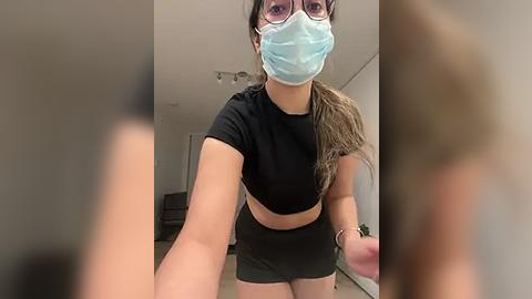 Media: Video of a young woman in a black crop top and shorts, wearing a blue surgical mask and glasses, taking a selfie indoors.