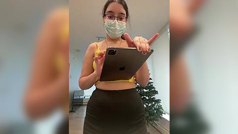 Media: Video of a young woman with fair skin, glasses, and a mask, holding a tablet in a bright, modern room. She wears a yellow crop top and dark high-waisted pants.