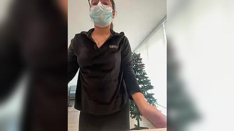 Media: Video of a woman in a black fleece jacket and mask, standing in a modern, brightly lit room with a Christmas tree in the background.