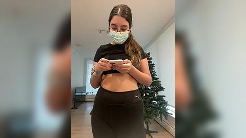 Media: Video of a young woman in a black crop top and pants, wearing a mask, holding a phone, standing in a modern, bright room with a Christmas tree.