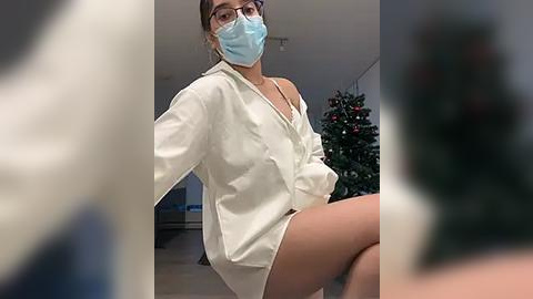 Media: Video of a young woman with light skin, wearing a blue mask, a white shirt, and no pants, seated on a chair in a room with a Christmas tree.