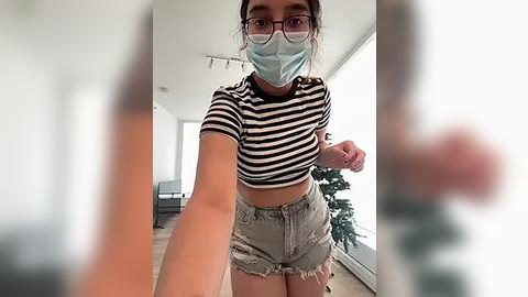 Media: Video of a young woman with fair skin, glasses, and a blue face mask, wearing a black-and-white striped crop top and ripped denim shorts. Background shows a modern living room with a Christmas tree.