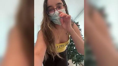 Media: Video of a young woman with long brown hair, wearing a yellow mask, black-rimmed glasses, and a black and yellow tank top. She's holding a piece of food to her mouth.