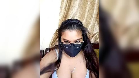 Media: Video of a woman with long dark hair, wearing black face mask, glasses, and light blue tank top, revealing cleavage. Background features gold curtains and blurred figures.