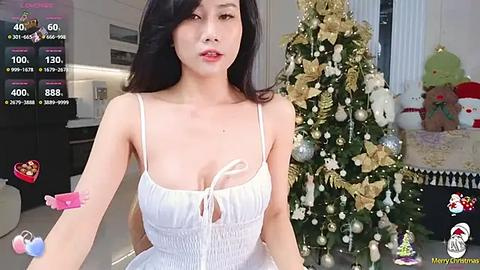 Media: Video of a young Asian woman with long black hair, wearing a white dress, standing in front of a decorated Christmas tree in a modern, bright room.