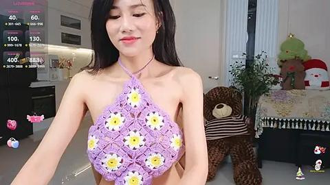 Media: A video of an East Asian woman with long black hair, wearing a crocheted purple halter top adorned with yellow daisy flowers. Background features a modern living room with plush toys, plants, and a plush teddy bear.