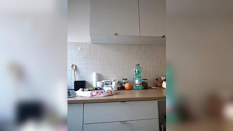 Media: Video of a modern kitchen with white cabinets, a light wood countertop, and a tiled backsplash. The counter holds a water bottle, oranges, a plate of cookies, and various utensils.